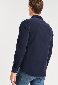 Marine corduroy overhemd, lange mouwen, knoopmanchetten, subtiele ribbeltextuur, gebogen zoom, gedragen met blauwe jeans. Geen zichtbare patronen of accenten.