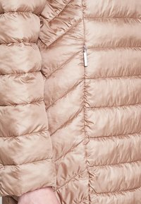 Beige pufferjack met een quilted chevronontwerp, een gladde textuur en een ritszak aan de zijkant. Lichtgewicht en geïsoleerd.