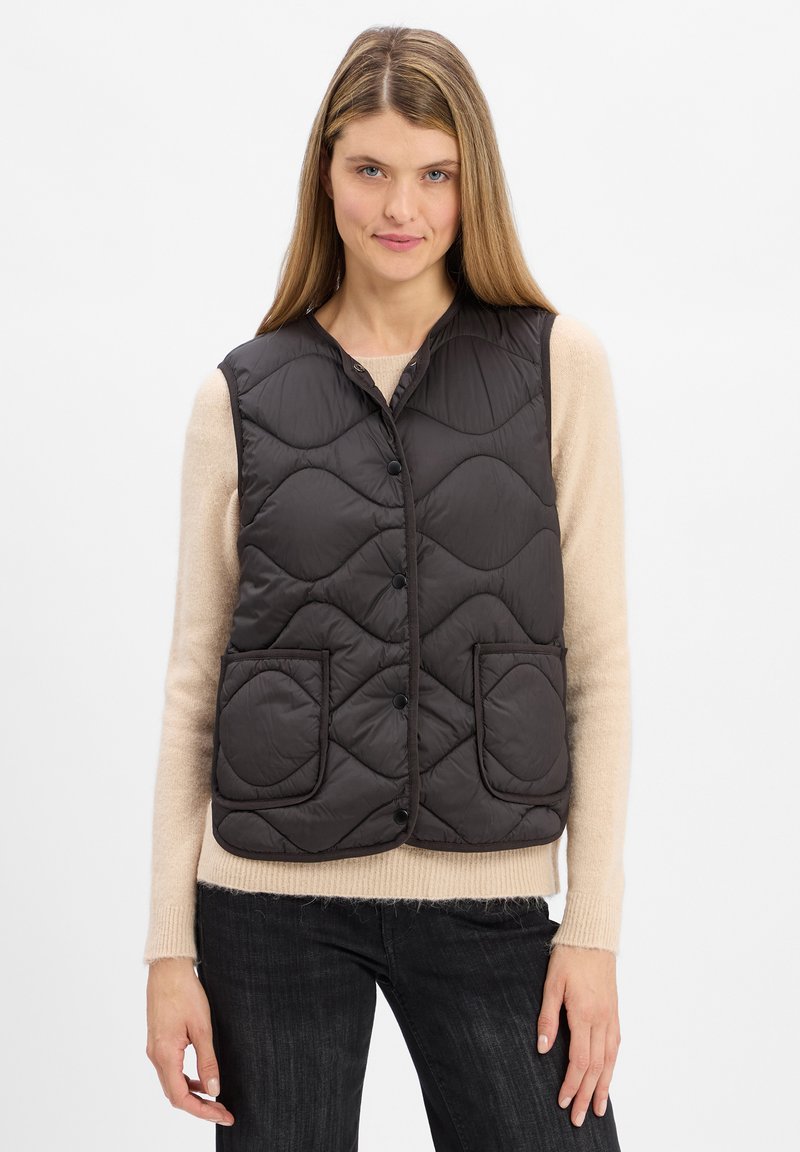 Gepolsterte dunkelbraune Weste mit Stehkragen, ausgestattet mit zwei runden Fronttaschen und Druckknöpfen. Getragen über einem hellen Pullover.