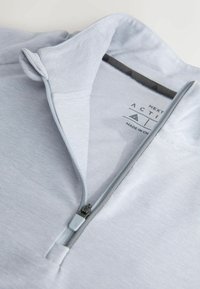 Gai pelēkā ceturtdaļas rāvējslēdzēja sporta krekls ar izcilu apkakli un redzamu melnu un pelēku rāvējslēdzēja vilkšanu, nolikts uz baltas fona.