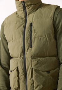 Oliven grøn puffer vest med høj krave, frontlynlås og to store påsyede lommer; tekstureret stof med kompakt design og sorte hardwaredetaljer.