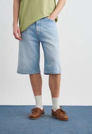 Άτομο που φοράει ανοιχτό μπλε τζιν σορτς μέχρι το γόνατο, πράσινο μπλουζάκι, λευκές κάλτσες και καφέ δερμάτινα παπούτσια με κορδόνια, στέκεται πάνω σε μπλε χαλί.