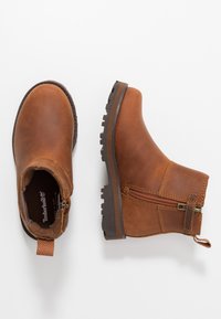Bottes cheville en cuir marron avec une fermeture éclair sur le côté, semelle en caoutchouc, et un dessus texturé avec des détails de couture et une languette.