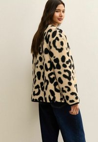 Woman with long dark hair wearing a beige and black leopard print fleece jacket and dark blue jeans, facing slightly sideways and smiling.