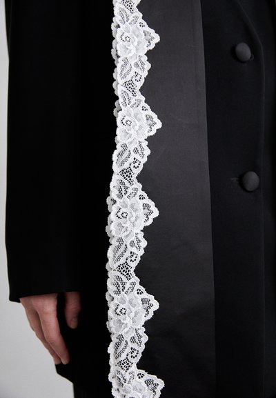 Zwarte stoffen jas met een delicate witte kantafwerking langs de rand, voorzien van bloemmotieven en een knoopdetail op de mouw.