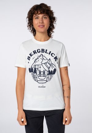 Frau mit lockigem Haar, die ein weißes T-Shirt mit einer Grafik eines Berges, eines Rucksacks und eines Hutes mit dem Text "BERGBLICK" und "Wandermädel" trägt.