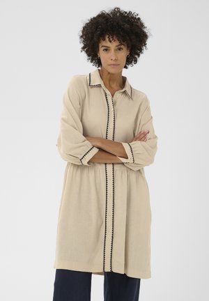 Kvinde med krøllet hår iført en beige knappet tunika med sort zigzagkant, armene krydset, stående mod en ensfarvet lys baggrund.