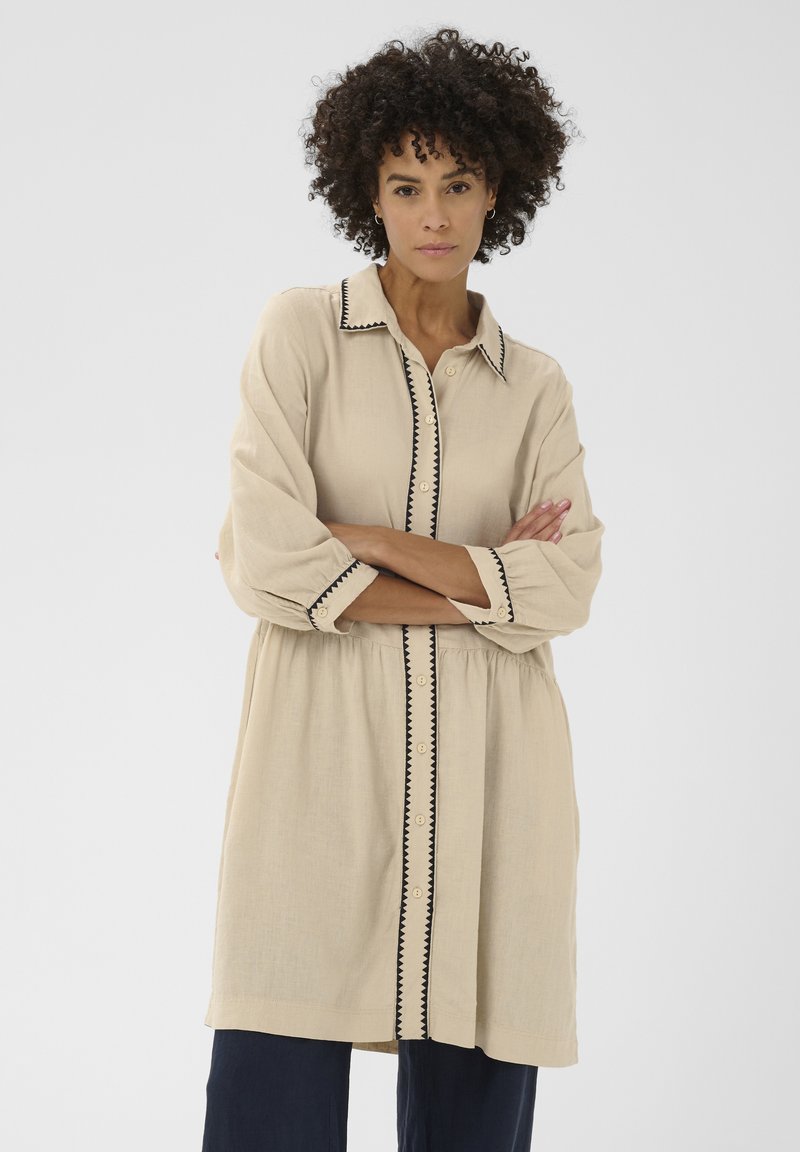 Femme aux cheveux bouclés portant une tunique beige boutonnée avec un bord en zigzag noir, bras croisés, debout devant un fond clair uni.