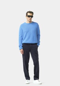 Helles blaues strukturiertes Sweatshirt, dunkle Cordhose, weiße Sneaker und schwarze Sonnenbrille. Schlichte Silhouette mit gerippten Bündchen und Saum.