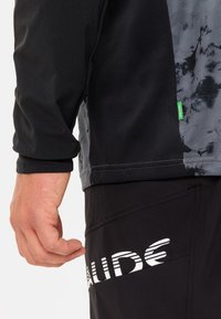 Langermet svart og grå treningsgenser med et teksturert mønster, med et grønt merkelapp, sammen med svarte shorts som har hvit merkevare.