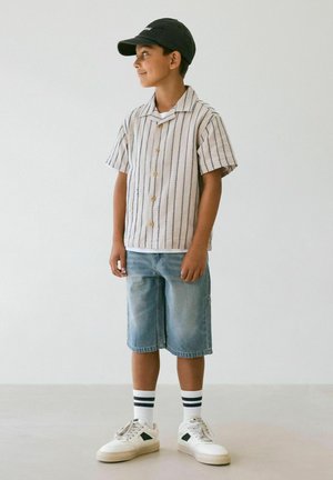 Ragazzo con berretto nero, camicia a righe a maniche corte con bottoni, pantaloncini di jeans al ginocchio, calzini bianchi con righe nere e scarpe da ginnastica bianche.