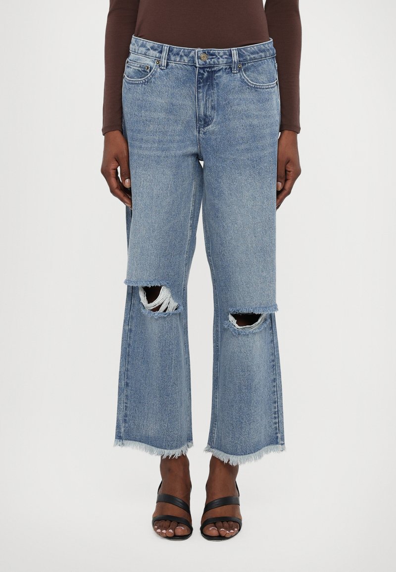 Jeans a gamba larga in denim blu chiaro con orli sfrangiati e ampie aperture sulle ginocchia. Design a cinque tasche con chiusura a bottone e passanti per cintura.