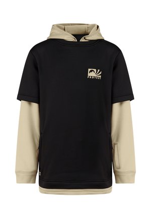 Teenager-Junge steht und trägt einen schwarzen kurzärmeligen Hoodie über einem beigefarbenen langärmeligen Shirt, beige Hose und schwarze Schuhe vor schlichtem Hintergrund.