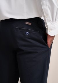 Zbliżenie na osobę w czarnych spodniach z tylną kieszenią zapinaną na guzik oraz białej koszuli z długim rękawem, ręka częściowo w kieszeni bocznej.