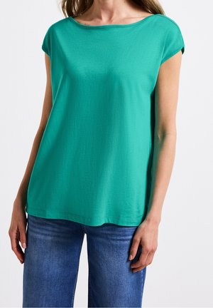 Kobieta w turkusowym topie z krótkim rękawem połączonym z niebieskimi jeansami na jednolitym białym tle.