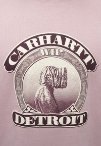 Svetlo vijolična majica s kratkimi rokavi, na kateri je grafika osebe v kapucni jakni. Črn napis "CARHARTT WIP" in "DETROIT" obkroža dizajn.
