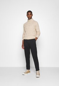 Suéter de punto beige de cuello alto con puños acanalados, combinado con pantalones tapered oscuros de color azul marino y zapatillas altas beige.