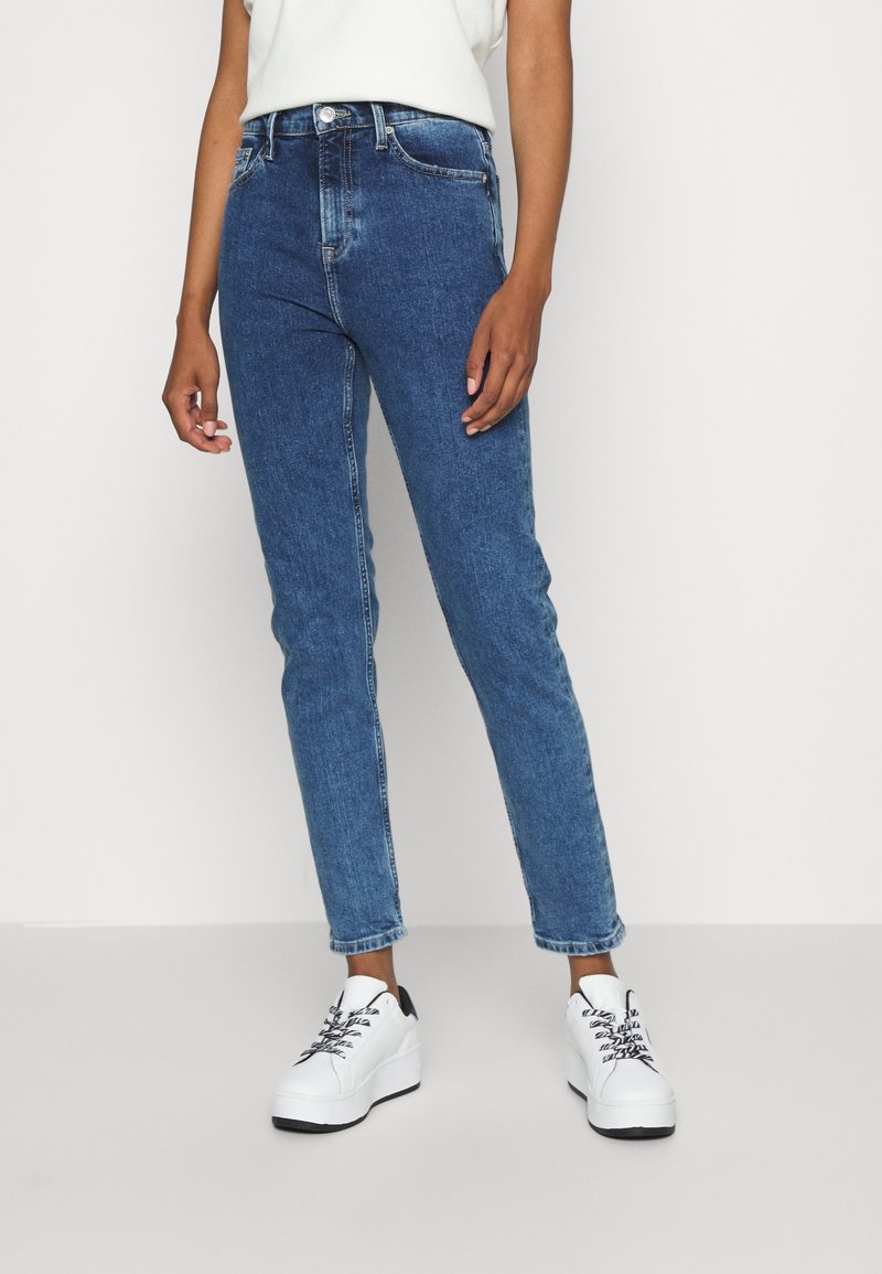 Personne portant un jean bleu taille haute, un haut blanc sans manches et des baskets blanches à plateforme, debout devant un fond uni clair.