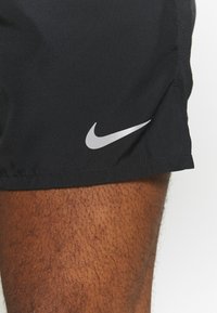 Mustat Nike-shortsit, joissa on teksturoitu kangas, hopeinen logo vasemmassa alakulmassa ja pieni halkio helman kohdalla.