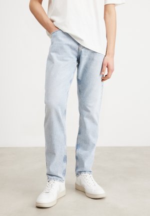 Personne portant un jean droit bleu clair, des baskets blanches et un t-shirt blanc, debout avec une main dans la poche.