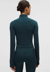 Kobieta z ciemnymi włosami związanymi w kok, ubrana w dopasowaną, ciemnoniebieską długą bluzę sportową i pasujące legginsy, przedstawiona z tyłu.