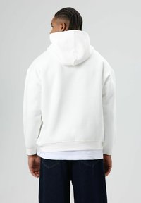 Witte capuchontrui met een losse pasvorm, geribde manchetten en zoom. Voorzien van een grote capuchon en een zachte textuur. Draagt als een laagje over een wit kledingstuk.