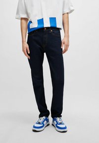 Jeans de mezclilla azul oscuro con un diseño ajustado, combinados con una camisa blanca que presenta un patrón geométrico azul en el dobladillo, y zapatillas deportivas azules.