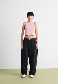 Jeune personne avec les cheveux courts et sombres, portant un crop top rose sans manches, un jean noir oversize et des baskets blanches sur un fond uni.