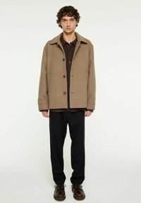 Brun ulljacka med krage, knappstängning och sidofickor, bärs över en mörk skjorta och kombineras med svarta byxor och skor.