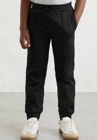 Czarne spodnie dresowe wykonane z miękkiego materiału, z bocznymi kieszeniami, elastycznym pasem ze sznurkiem oraz zwężanymi mankietami. Zestawione z białymi butami sportowymi.