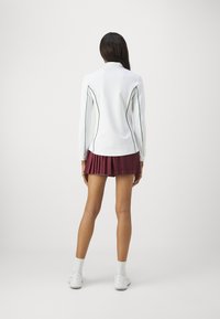 Vit långärmad topp med gröna sidsömmar, kombinerad med en vinröd plisserad kjol. Outfiten har en sportig design, lämplig för aktiv klädsel.