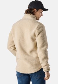 Beige fleece pullover with a high collar, textured fabric, and slim fit. Long sleeves and slightly curved hem. Worn over blue jeans.