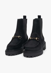 Černé semišové chelsea boty s robustní černou platformovou podrážkou, zlatými kovovými akcenty a texturovaným povrchem svršku.