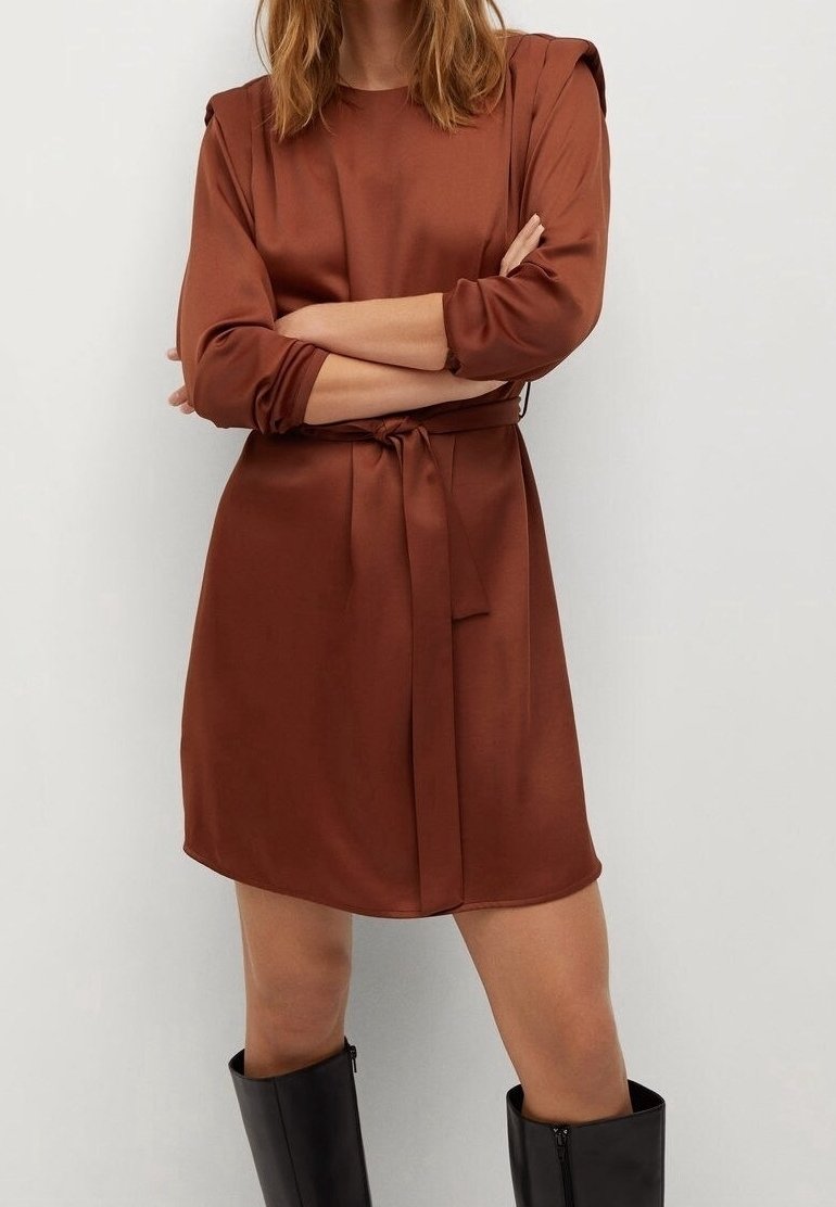 Femme portant une robe marron mi-longue avec des manches longues et une ceinture à la taille, associée à des bottes noires montant jusqu'aux genoux, les bras croisés.