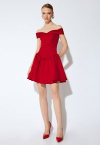 Robe rouge décollet�ée avec un corsage ajusté et une jupe évasée. Texture de tissu lisse, longueur genoux, assortie à des talons hauts rouges.