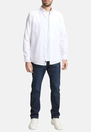 Homme portant une chemise blanche boutonnée par-dessus un t-shirt blanc, un jean bleu foncé et des baskets blanches, debout devant un fond uni.