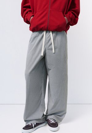 Person trägt roten Kapuzenpullover mit Reißverschluss, lockere graue karierte Hose mit Kordelzug und dunkle Sneaker mit weißen Sohlen vor neutralem Hintergrund.