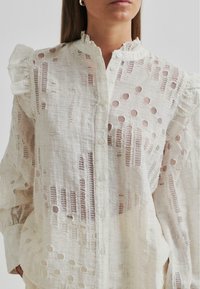Light linen blouse with ruffled shoulders, white with sheer cut-out patterns. Features a button-down front and long sleeves.