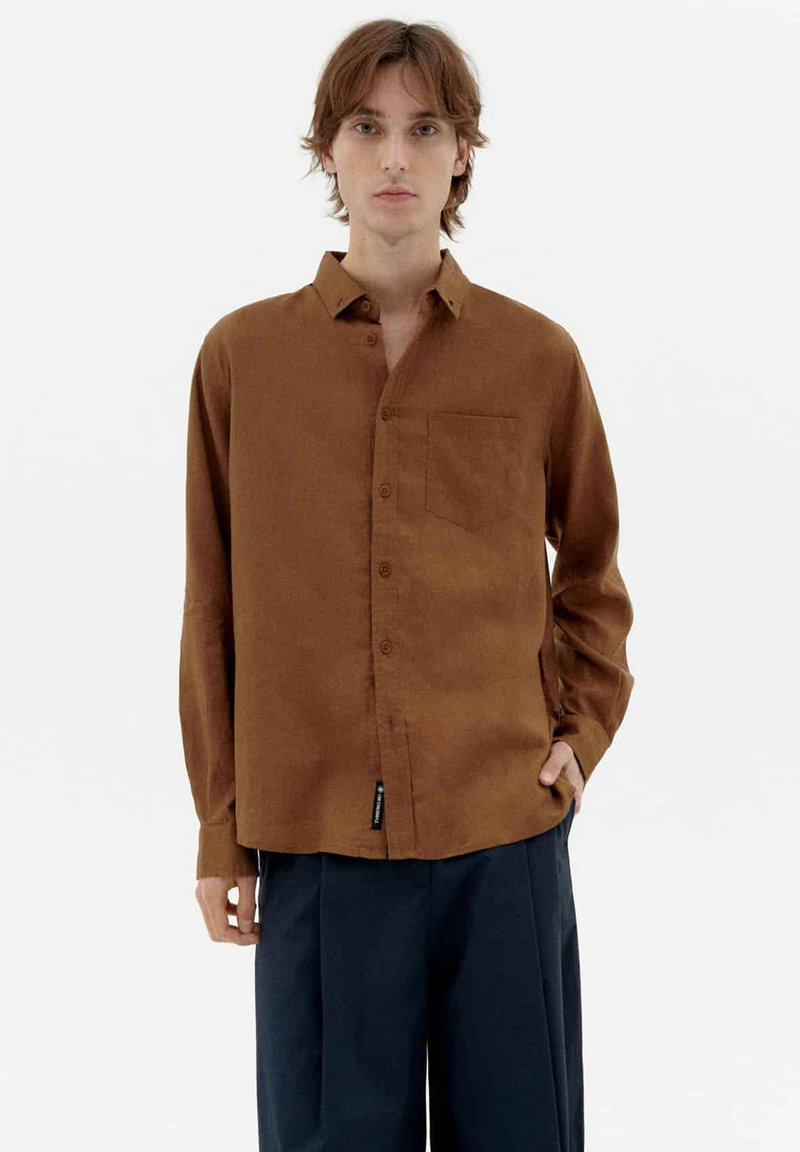Brown long-sleeve button-up shirt with a patch pocket, made of fabric with a smooth texture. Paired with dark navy trousers.