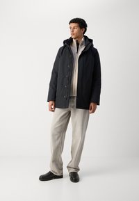 Chaqueta negra aislante con capucha, con botones a presión y una capa interior con cremallera, combinada con pantalones vaqueros holgados gris claro y zapatos negros.