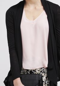Schwarze leichte Strickjacke über einem pinkfarbenen V-Ausschnitt-Oberteil. Das Outfit wird mit floral gemusterten Hosen und einer schwarzen Clutch-Tasche kombiniert.