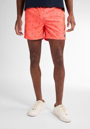 Korallenfarbene Badeshorts mit kleinen schwarzen Mustern, elastischem Bund und Seitentaschen. Getragen mit weißen Sneakers vor einem neutralen Hintergrund.