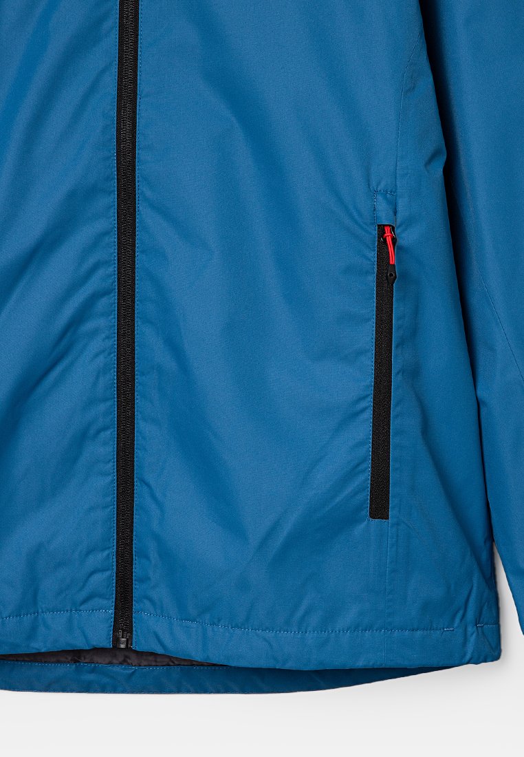 Veste bleue avec une texture lisse, dotée d'une fermeture éclair noire et d'une poche zippée avec des accents rouges. Design simple et contemporain.