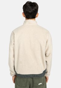 Kurtka polarowa w kolorze kremowym z wysokim kołnierzem, z ciemnozielonym wykończeniem. Miękka tekstura, luźny krój i długie rękawy.