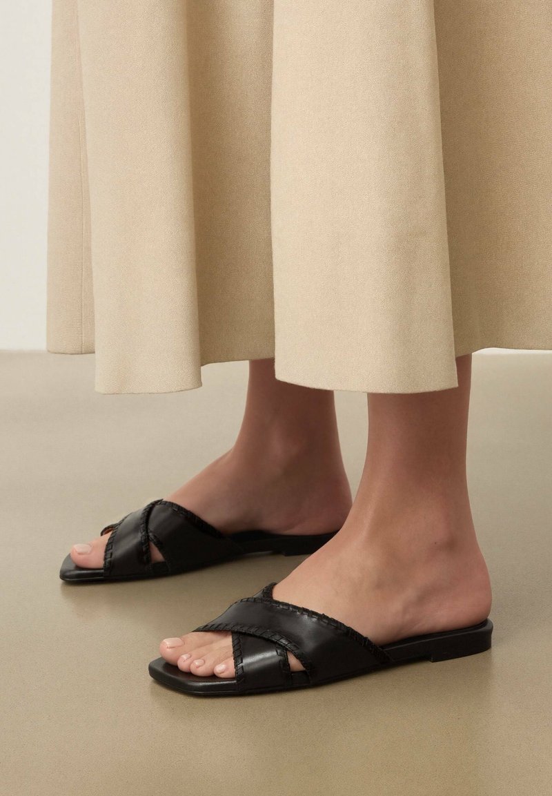 Pieds chaussés de sandales plates en cuir noir avec des brides tressées croisées, accompagnées d'un pantalon beige à jambes larges, ourlé au-dessus des chevilles.