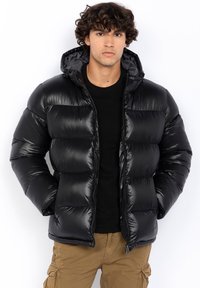 Doudoune noire avec une finition brillante, fermeture éclair à l'avant et capuche. Présente un motif matelassé et des poignets élastiques. Portée sur une chemise noire.