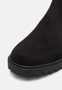 Gros plan d'une botte en daim noir avec un bout arrondi et une semelle épaisse en caoutchouc à crampons, sur fond blanc.