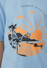 Lichtblauw katoenen t-shirt met een cirkelvormige afbeelding van zwarte palmbomen, een oranje zon en golven, en de tekst "DE ZOMER BELLING."