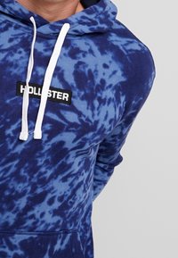Hoodie bleu tie-dye avec des cordons blancs, comportant un patch noir avec le nom de la marque "HOLLISTER". Fabriqué en tissu doux, coupe décontractée.