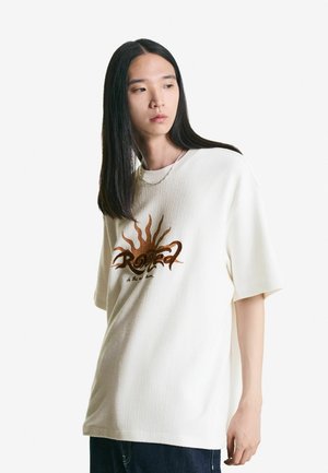 Hvid ribbet t-shirt med et brunt grafisk design af en sol og tekst. T-shirten har en afslappet pasform og korte ærmer.